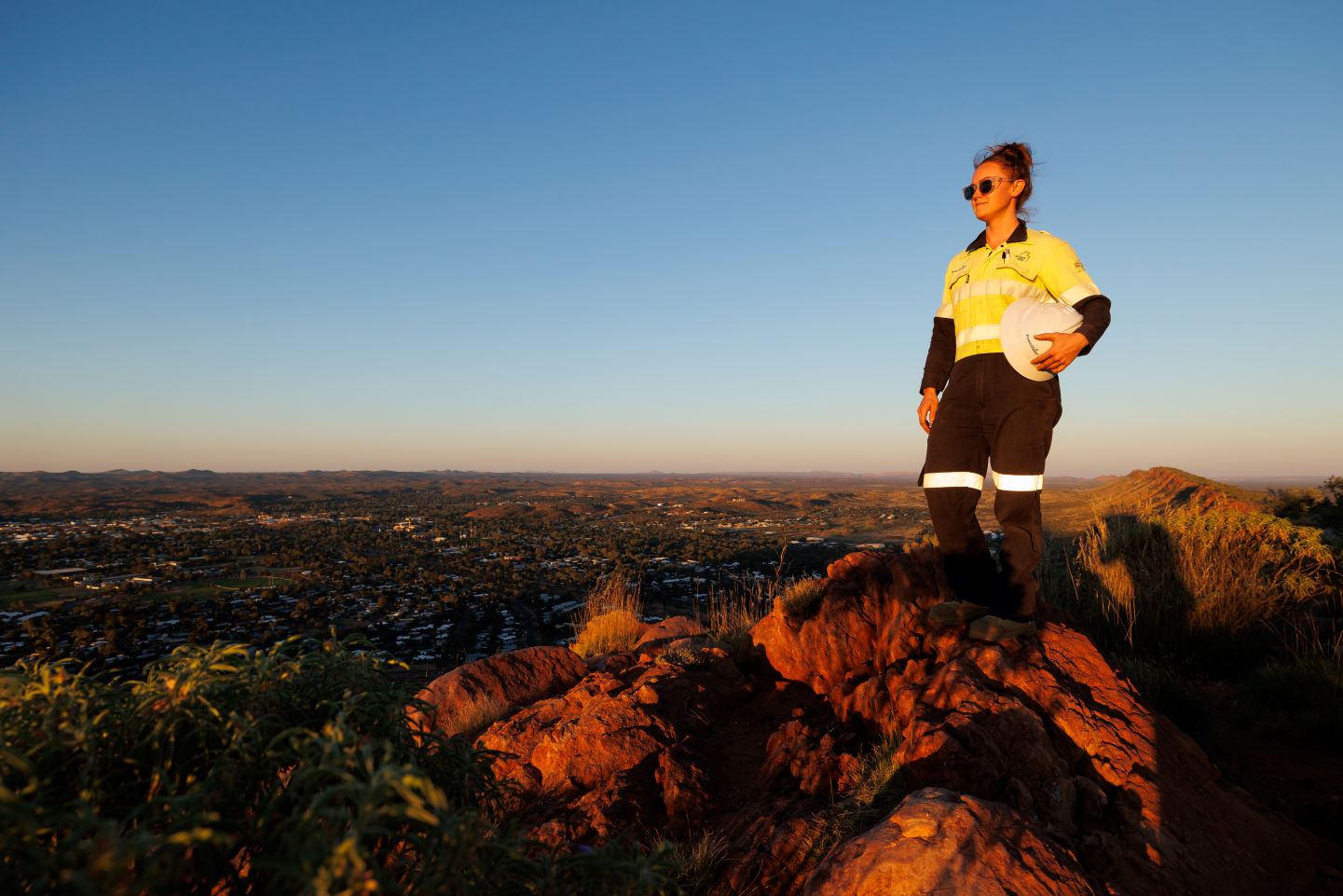 Smiling Power and Water employee wearing a yellow high-visibility in personal protective equipment uniform wearing a brown hat, holding a tablet while standing on a hill overlooking a rural landscape at sunset.
